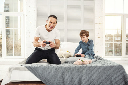 Playing games. Handsome exuberant well-built father smiling and playing games with his son and they holding remote controls