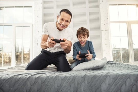 Relaxing at home. Good-looking content well-built father smiling and playing games with his son and they holding remote controls