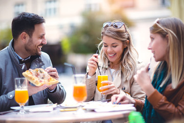 Group of three friends using phone in outdoor cafe on sunny day, eating pizza