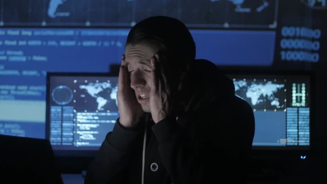 Male geek hacker overworking at computer and suffers from a headache in cyber security center filled with display screens.