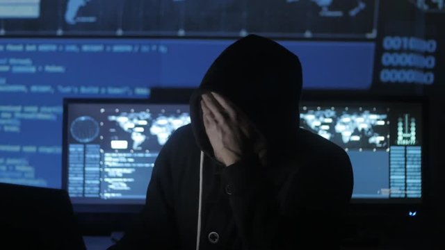 Man geek hacker in hood overworking at computer and suffers from a headache in cyber security center filled with display screens.