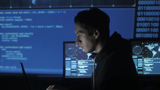 Geek Hacker programmer is working on computer in cyber security center filled with display screens.