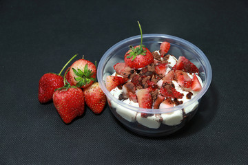 Home made cake and white buttercream, topping by Strawberries cut into pieces with cocoa powder in the round plastic cup. And Strawberry on the black floor.