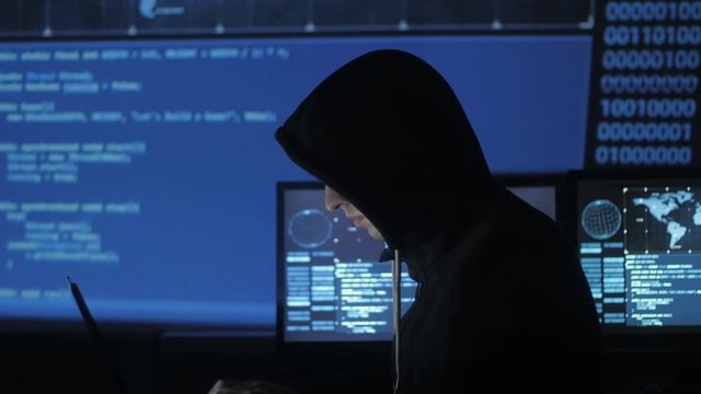 Man geek hacker in hood working at computer in cyber security center filled with display screens.