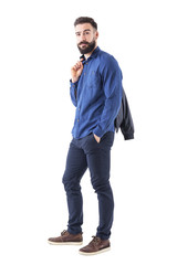Fototapeta premium Side view of young bearded man in blue shirt carrying bomber jacket over the shoulder looking at camera. Fully body isolated on white background. 