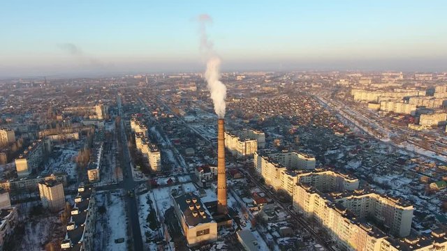 A bird`s eye view of a high cooling tube with dense swhite moke between multistoried buildings at sunset in winter. The drone flies up 
