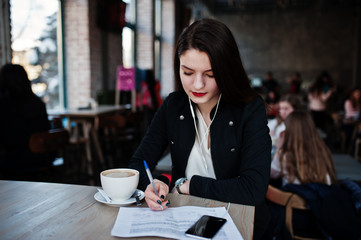 Brunette girl sitting on cafe with cup of cappuccino, listening music on headphones and write some documents.