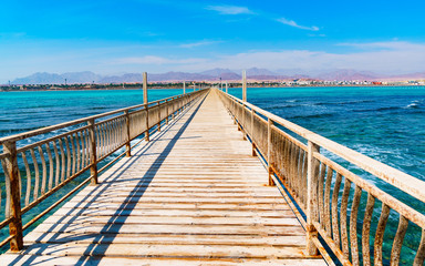 Abandoned sea pier, leading into the distance. Rusty railing. Old wooden boards. View of the mountain shore. Coudy blue sky. Sunny day.