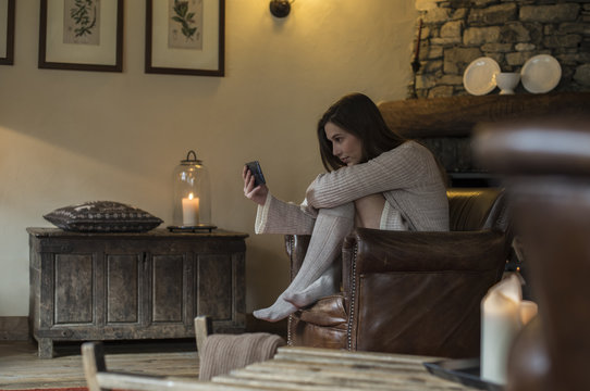 young woman in casual dressing at home relaxing on armchair by using smartphone to browse internet or looking at photos during cold autumn or winter season