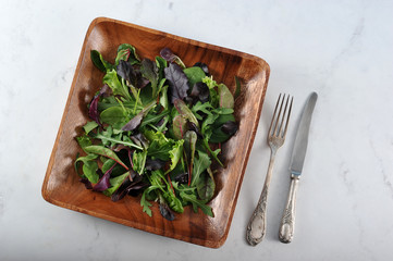 A mixed of a variety of lettuce leaves in a wooden dish. Rukkola, maingold, corn, lettuce. Next to the dish are cutlery. Light background. View from above. Close-up. Macro photography.