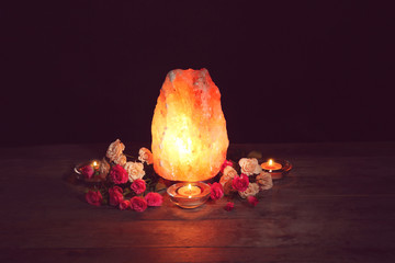 Himalayan salt lamp, candles and flowers on table against dark background