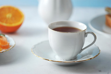 A cup of black tea and a saucer. In the frame there is a plate, a teapot and half an orange. Light background. Close-up. Macro photography.