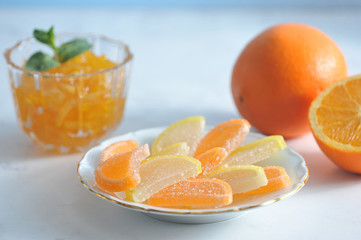 Marmalade in the form of orange and lemon slices on a plate and orange jam in a vase. The jam is decorated with mint leaves. Next to an orange and half an orange. Light background. Close-up.
