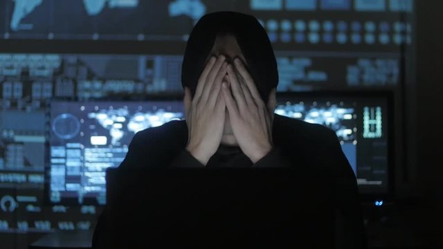 Man geek hacker in hood overworking at computer and suffers from a headache in cyber security center filled with display screens.