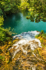 Naklejka na ściany i meble Widok z nad wodospadu. Woda wpadająca do zielonego jeziora. Jezioro wśród drzew w paku Krka w Chorwacji.