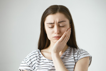 Portrait of unhappy depressed young Caucasian brunette woman closing eyes in frustration and pressing hand against cheek, trying to soothe pain while suffering from toothache, having tooth decay
