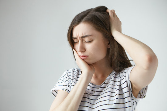 People, sickness and bad health condition concept. Picture of unhappy frustrated young brunette woman closing eyes, touching cheek and back of her head while suffering from terrible tooth ache