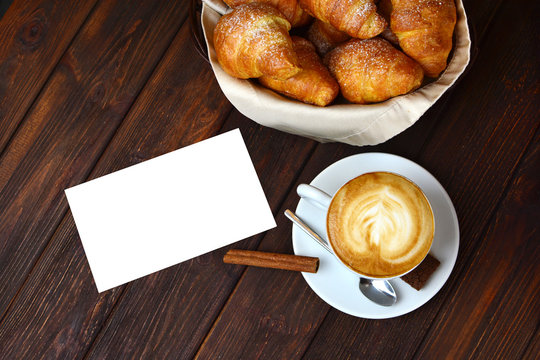 Background with a cappuccino, empty white paper and croissants on a wooden textured table.