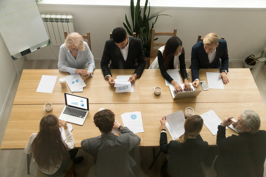 Multiracial young and old business people sitting at conference table meeting in office, diverse team group working together with devices discussing documents, teamwork concept, top view from above