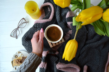 Stylish spring still life. Sweater, a cup of coffee in female hand, napoleon cake, yellow tulips, eyewear on the white wooden background. Beautiful morning flat lay.