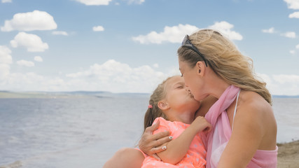 Little cute girl baby and young mother at beach. Mom sits on the shore, and her daughter is coming from behind. The kid hugs and kisses her mother. Mother and her child.