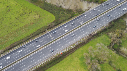 Aerial view of a stretch of highway in the Italian countryside. The road is divided into many lanes in each direction. There are cars and trucks on the street.Around the road there are trees and grass