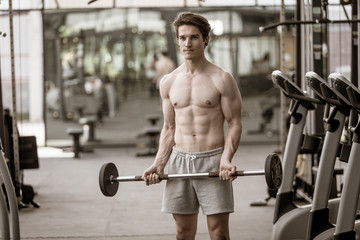 A shirtless young muscular man working out in gym doing exercises with equipment in fitness center.