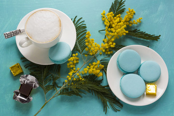 A cup of cappuccino coffee with mint spiked macaroni and chocolate on a mint-blue table with a branch of a blossoming mimosa. The concept of spring coffee, the morning of the women's holiday.