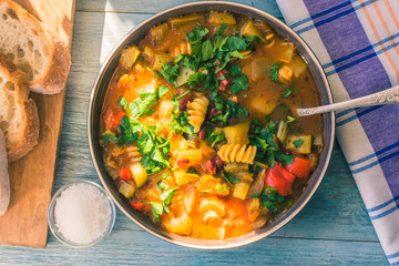 Traditional minestrone soup with pasta and chopped green parsley in a plate on a rustic wooden table close-up with a napkin, sunny day - top view, copy space for a recipe