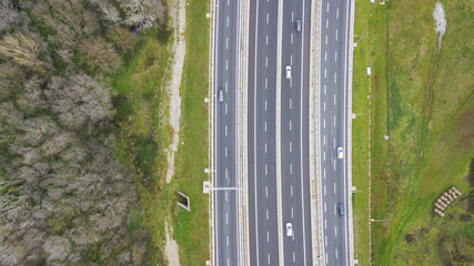 Aerial view of a stretch of highway in the Italian countryside. The road is divided into many lanes in each direction. There are cars and trucks on the street.Around the road there are trees and grass