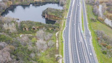 Aerial view of a stretch of highway in the Italian countryside. The road is divided into many lanes in each direction. There are cars and trucks on the street.Around the road there are trees and grass