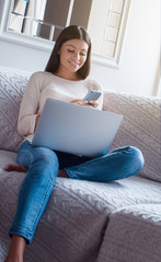 Working from home. Beautiful young woman using her smart phone with smile while sitting on the sofa with laptop on her knees in home interior