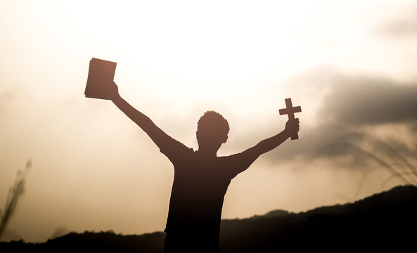 Human holding Bible book and christian cross with light sunset background