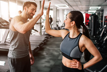 A beautiful girl and her well-built boyfriend are greeting each other with a high-five. They are happy to see each othr in the gym. Young people are ready to start their workout. - obrazy, fototapety, plakaty