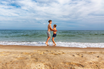 Obraz na płótnie Canvas Two teenagers: a girl and a boy with blond hair, dressed in white T-shirts and shorts jumping up holding hands on a sea beach. Copy space.