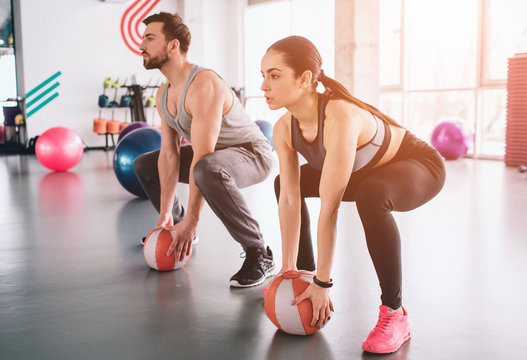 Strong guy and a slim girl are sitting in a position, ready to do squats. They have put their hands on balls ready to take them and hold it over their hands in any time.