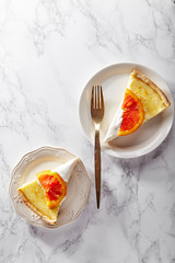 Slices of soft cheesecake with caramelized red oranges and whipped cream served on plates of different size and from on white marble table