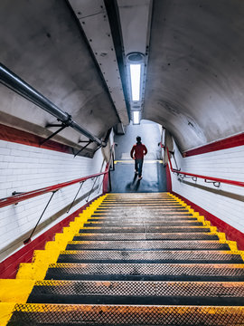 Young man in the red jacket rushing down the stairs of the Tube underpass. White ceramic wall tiles and metal staircase with red handrail at Chalk Farm Underground Station.