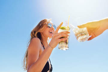 Smiling pretty woman with long hair in swimsuit and sunglasses, drinking a cocktail, toasting, celebrating something with friend, spending vacation on the beach, at sunny day. Bottom view