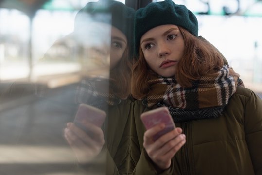 Thoughtful woman leaning on glass wall while holding smartphone
