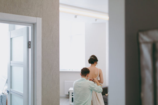 Morning preparation newlyweds for the wedding. A young guy in a light shirt fastens exquisite wedding dress for a brunette bride with a beautiful hairstyle and decoration in her hair in the room.