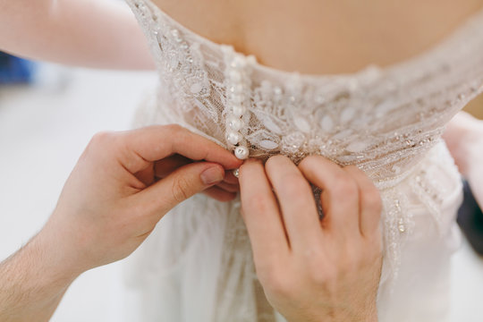 Close up Morning preparation newlyweds for the wedding. Men's hands button up elegant lace delicate ivory wedding dress with beads at the waist for bride. Wedding wear, accessories