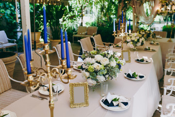 Beautiful and exquisite decoration of the wedding celebration in in the middle of a green garden. Banquet served table with a beige tablecloth, plates and candlesticks with blue candles under cover