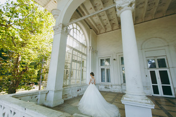 Obraz na płótnie Canvas Beautiful wedding photosession. Young bride in a white lace dress with a long plume with an exquisite hairstyle in vintage interior on the veranda of an old house with columns near the garden