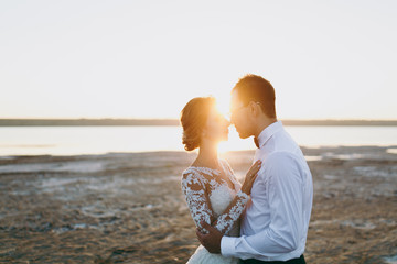 Beautiful wedding photosession. Handsome unshaved groom in a black trousers and young cute bride in white lace pattern dress with exquisite hairstyle on walk along the coastline near the sea sunset