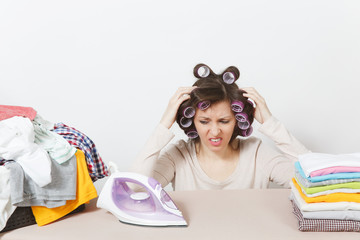 Distressed housewife with curlers on hair in light clothes ironing family clothing on ironing board...