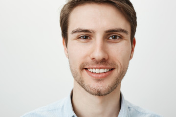 Fototapeta premium Close-up portrait of attractive confident adult male with bristle smiling broadly while looking at camera, standing against gray background. Calm and friendly office worker explains chores to newbie