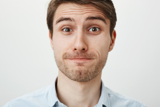 Close-up portrait of handsome clueless caucasian man with bristle standing with tight smile and lifting eyebrows, feeling awkward and embarrassed while standing against gray background.