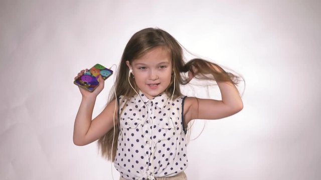 Child with earflaps dancing at studio background. The girl listens to music on the smartphone. The kid has loose long hair. Shooted on a gray white background.