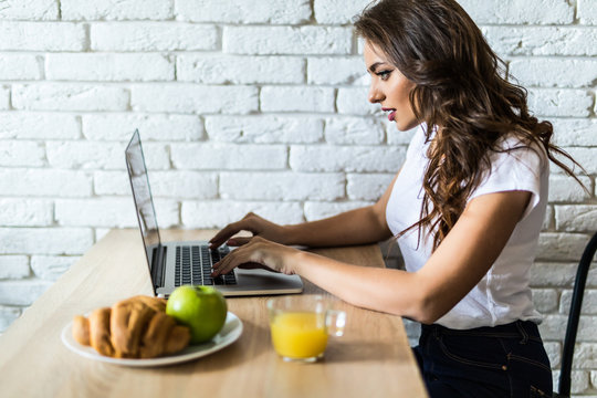 Young cheerful woman using on laptop computer and eating fruits in the kitchen in the morning